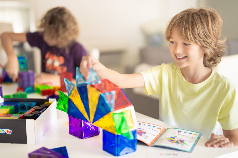 in Junge baut mit farbenfrohen Magneo Magnetbausteinen, während ein weiteres Kind im Hintergrund spielt. Kreatives, stabiles Bauen mit hochwertigem STEM Spielzeug von Magneo.