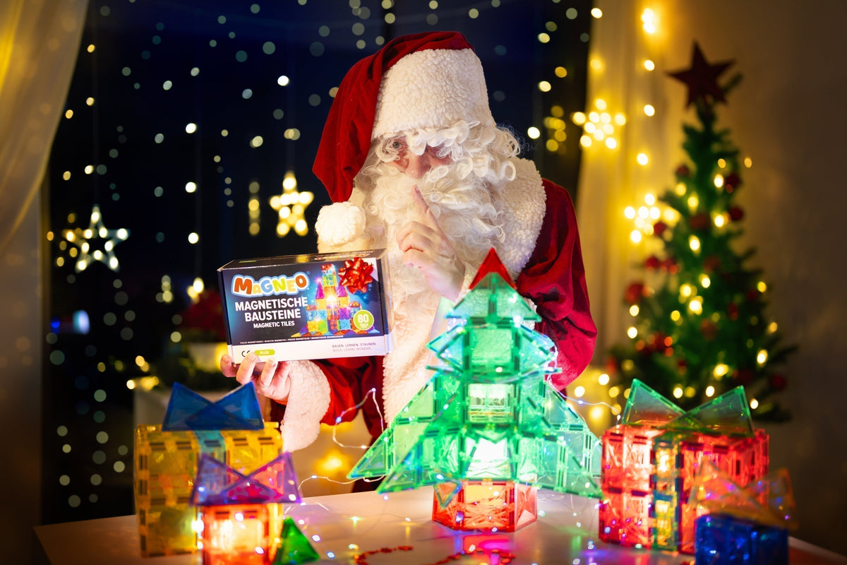 Weihnachtsszene mit einem verkleideten Weihnachtsmann, der die Magneo 80-Teile Box hält, umgeben von magnetischen Baustein-Bauten wie einem leuchtenden Weihnachtsbaum und Geschenkboxen.